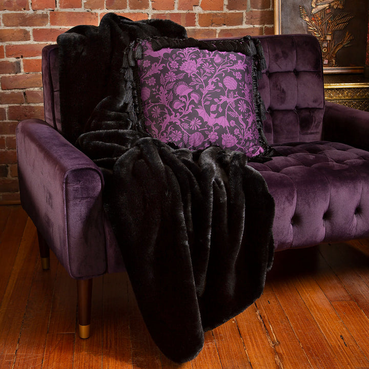 Black faux fur blanket draped over a purple velvet couch with a gothic botanical patterned throw pillow by Sin in Linen.