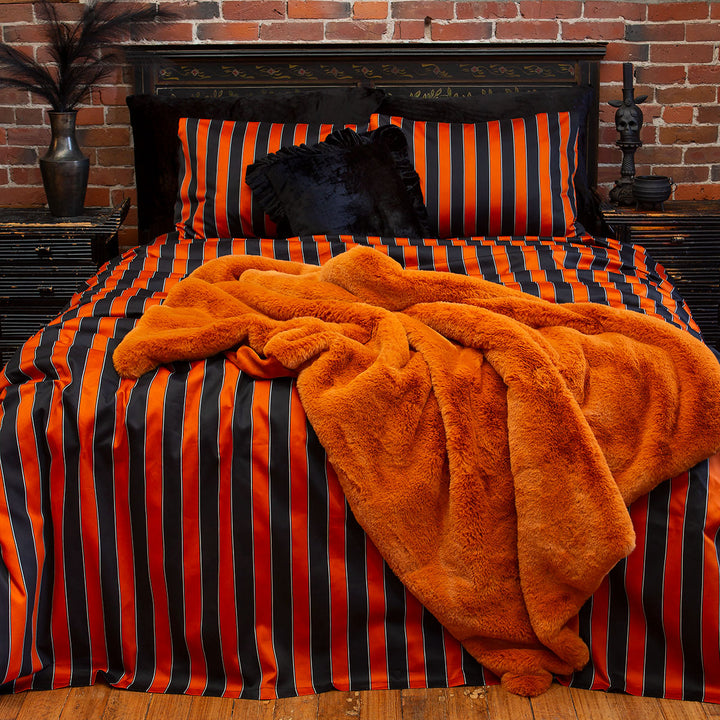 Pumpkin orange and black striped bedding on a bed. Inspired by vintage circus, gothic style for a Halloween all year round look. On the bed is a pumpkin orange fur blanket. 