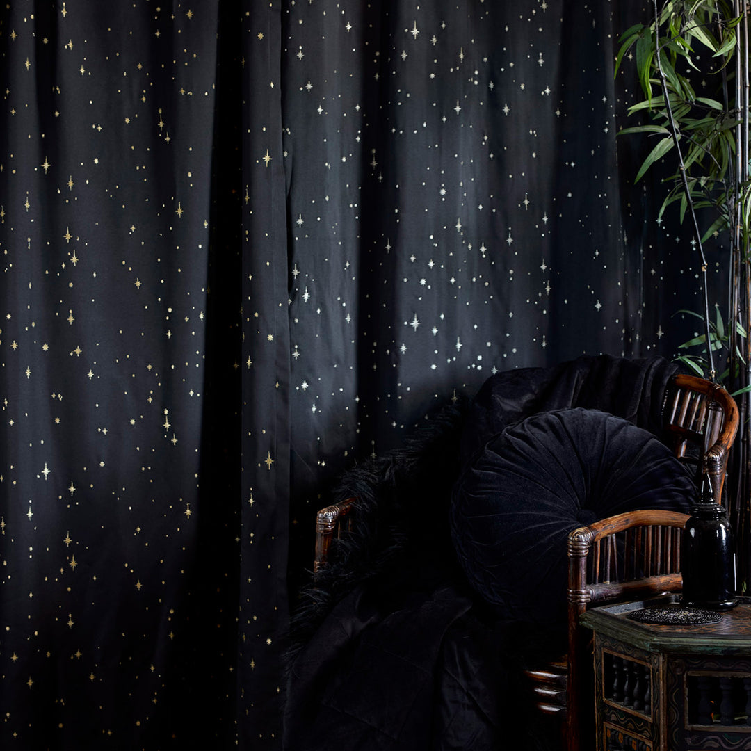 Sin in Linen's Night Sky Embroidered Curtain in Black with gold embroidered stars. Set in a lifestyle photo with a chair with velvet blankets and pillows. 
