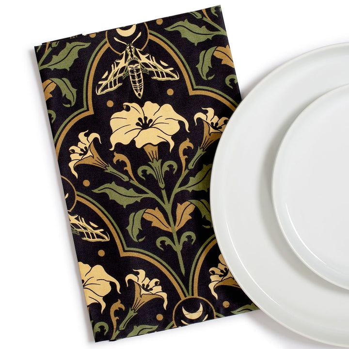 Datura Deco dinner napkins by Sin in Linen on a white background with a dinner plate. Gothic Botanical, witchy table linens featuring datura blooms, moths, and moons. 