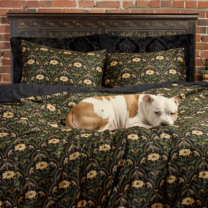 Gothic Botanical Art Deco Bedding with the Datura Deco print featuring moths, gilded accents, and blooming florals. On the bed are also velvet pillowcases and a sleeping puppy.