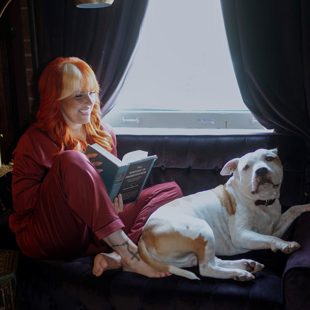 Woman with red hair reading a book next to a white dog on a couch wearing burgundy bamboo pajamas