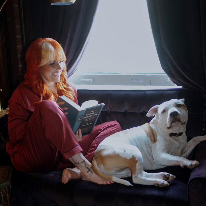 Woman with red hair reading a book next to a white dog on a couch wearing burgundy bamboo pajamas
