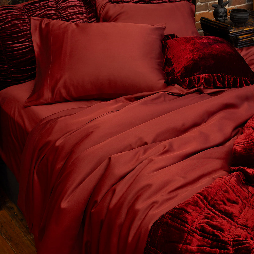 Oxblood burgundy red bamboo bed sheets on a bed with cozy velvet accent pillows and throw blanket. 