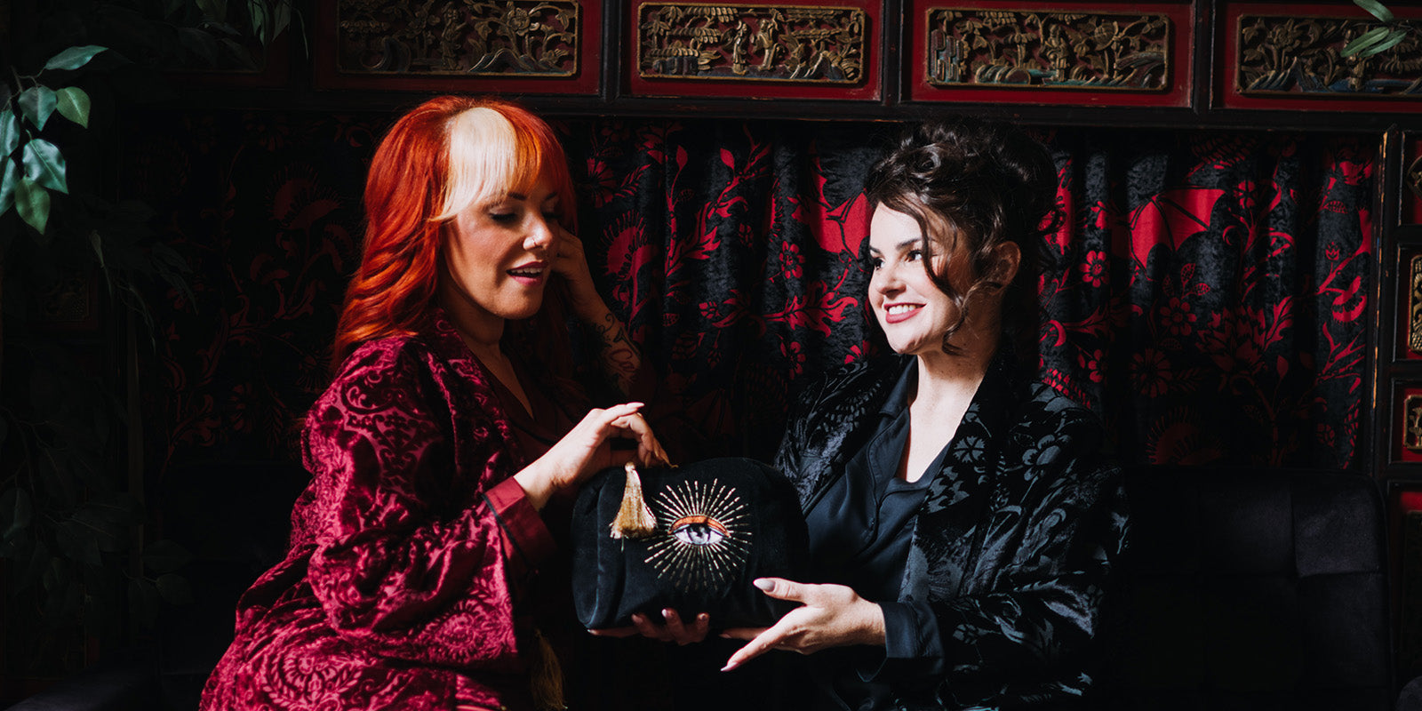 Two people holding a velvet toiletry bag with an embroidered eye design. They are wearing luxury bamboo pajamas and velvet robes by Sin in Linen.