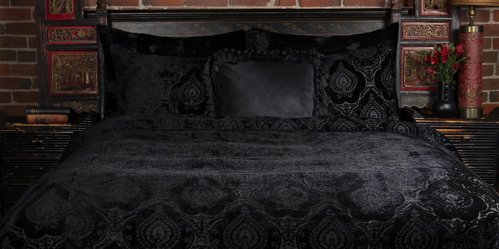 Black damask velvet bedspread on a bed with decorative pillows against a brick wall.