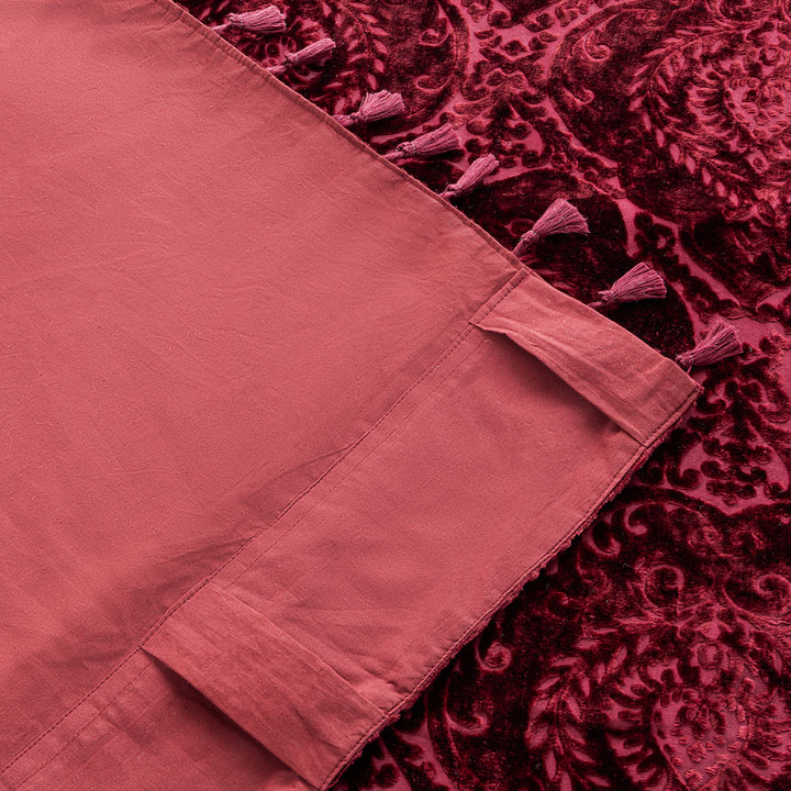Burgundy red damask flocked velvet curtain with a tasseled edge. The corner of the curtain is flipped up to show the cotton backing.