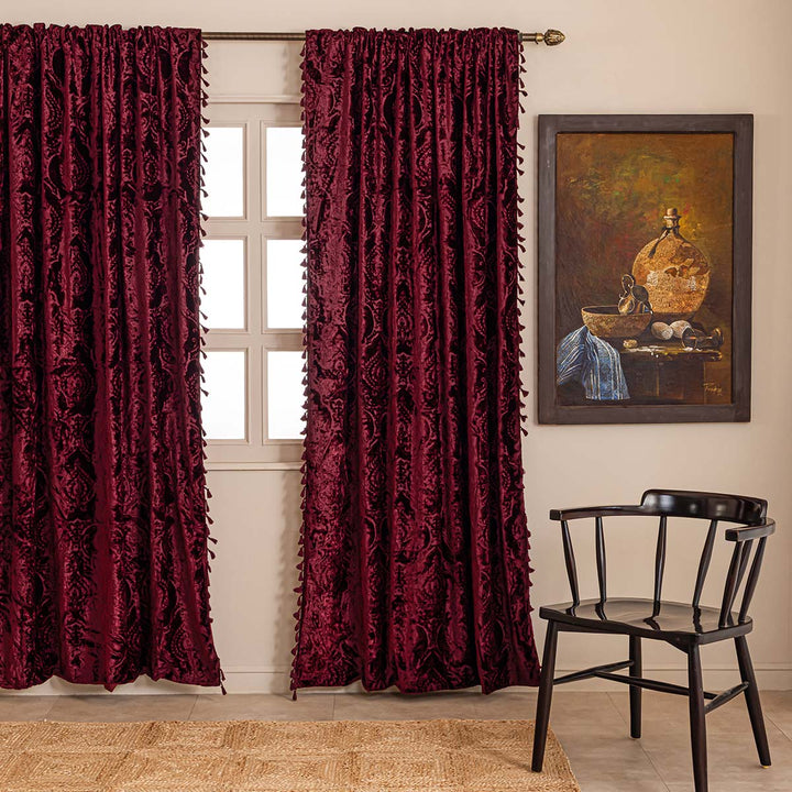 Burgundy red damask flocked velvet curtain with a tasseled edge in a room with a chair and painting.
