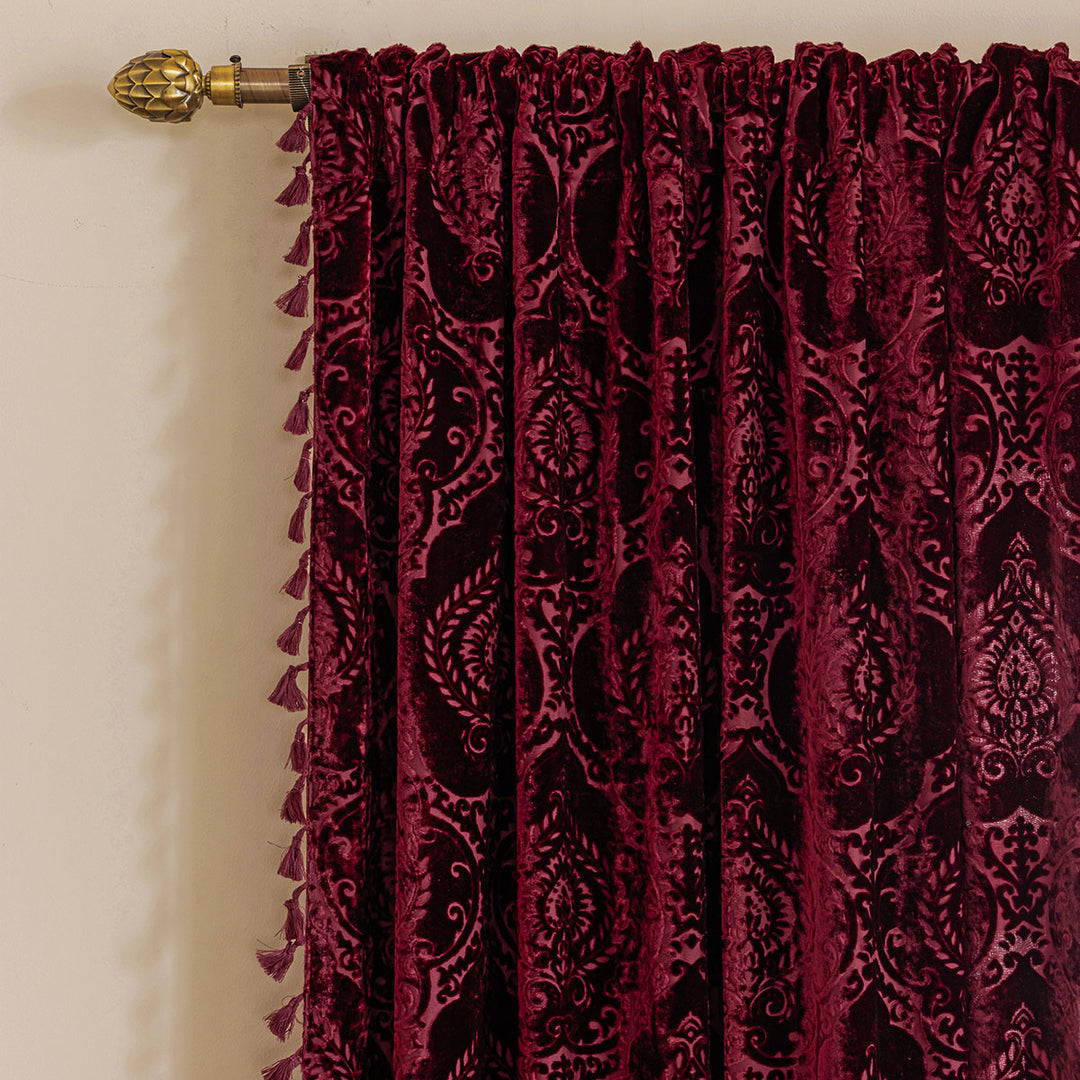 Burgundy red damask flocked velvet curtain with a tasseled edge on a curtain rod.