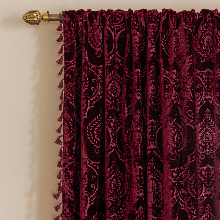 Burgundy red damask flocked velvet curtain with a tasseled edge on a curtain rod.
