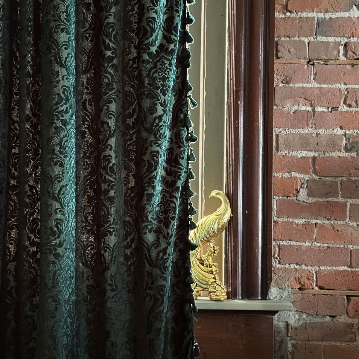 A section of emerald green velvet curtain with a damask pattern, tasseled edges, rod pocket, placed in front of a window with a brick wall in the background.